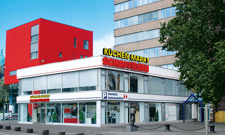 Das Bild zeigt den „Küchen-Markt Schaffrath“ in einem modernen zweistöckigen Gebäude mit einer weißen Fassade. Auf dem Dach befindet sich der Schriftzug „KÜCHEN-MARKT“ in gelben Buchstaben und „SCHAFFRATH“ in großen roten Buchstaben. Die untere Etage des Gebäudes hat große Glasfronten, durch die der Innenraum sichtbar ist, und bietet Einblick in die ausgestellten Produkte. Rechts neben dem Gebäude steht ein Baum, und im Hintergrund sind weitere moderne Gebäude zu erkennen, darunter ein auffälliges rotes Haus und ein mehrstöckiges Bürogebäude in Beige. Vor dem Küchen-Markt gibt es einen gepflasterten Bürgersteig mit Pollern, der Zugang zum Geschäft bietet. Der Himmel ist strahlend blau und vermittelt eine freundliche und einladende Atmosphäre.